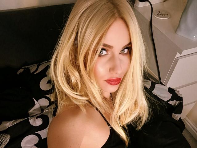 TinaBella has blonde hair, red lipstick, and is looking at the camera with a slight smile, showing her bare shoulders and black top. Her tits are partially visible.