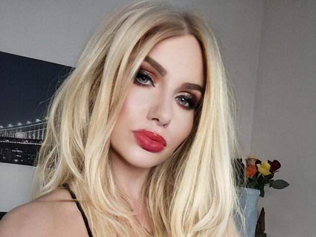 TinaBella with blonde hair, red lipstick, and smoky eye makeup, staring into camera, showing off her tits, looking hot. Background includes a blurry photo and flowers.