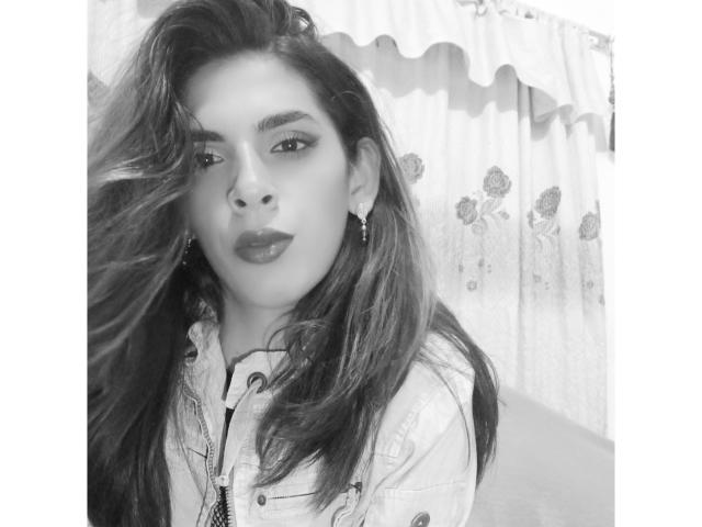 KataleyaHoty with long, wavy hair, wearing a denim jacket, dark lipstick, and earrings, looking seductively to the side, her ass slightly visible in the background. Black-and-white photo.