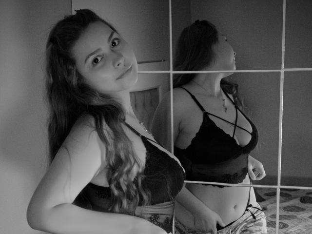 Fotografia in bianco e nero di RouseDawson con lunghi capelli ondulati, che indossa un reggiseno e dei pantaloni di pizzo, in piedi di fronte a uno specchio, mostrando il suo seno e il suo riflesso..