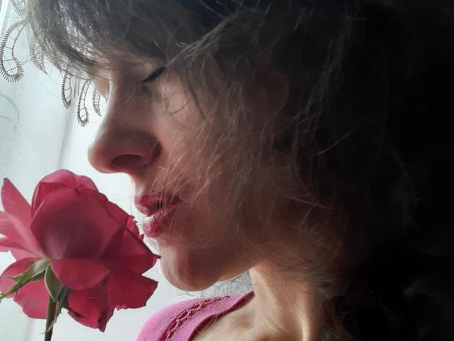 Le visage d'Orshiti en profil, tenant une rose rouge près de ses lèvres, avec des cheveux bruns foncés ondulés, vêtue de rose, lumière douce et légèrement ombragée, le nez et les lèvres d'Orshiti sont proéminents..