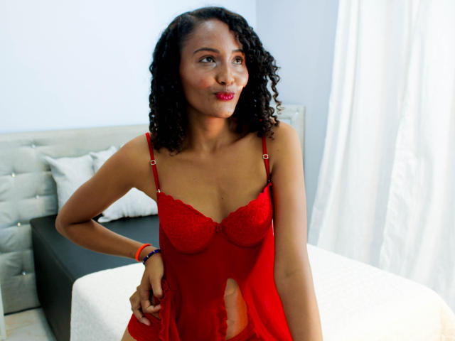 MeganGil with curly black hair, red lace lingerie, and red lipstick, standing in a bedroom, looking to the side, hand on hip, small red butt, medium tits, and red nipples.