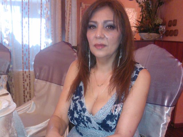 SxyVivian has brown hair, dark makeup, and a blue patterned dress with a low neckline showing her boobs. She sits with a purple-covered chair and a glass on the table.