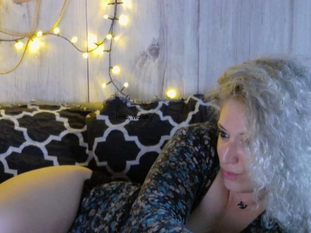 SassyKate with curly blonde hair, lying on a black and white patterned couch, wearing a floral dress, looking at the camera, with yellow string lights in the background.