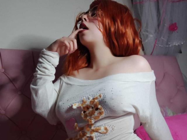 SummerTy, con cabello rojo, gafas y una blusa blanca con un hombro descubierto, chupándose el dedo, con la lengua afuera, con los pezones visibles, sentada en un sofá rosa, mirando hacia arriba..