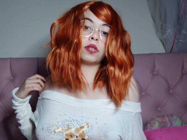SummerTy, com cabelo ondulado laranja, usando óculos e um suéter branco com um ombro de fora, com uma estampa de animal fofa, olhando para a câmera com batom rosa..