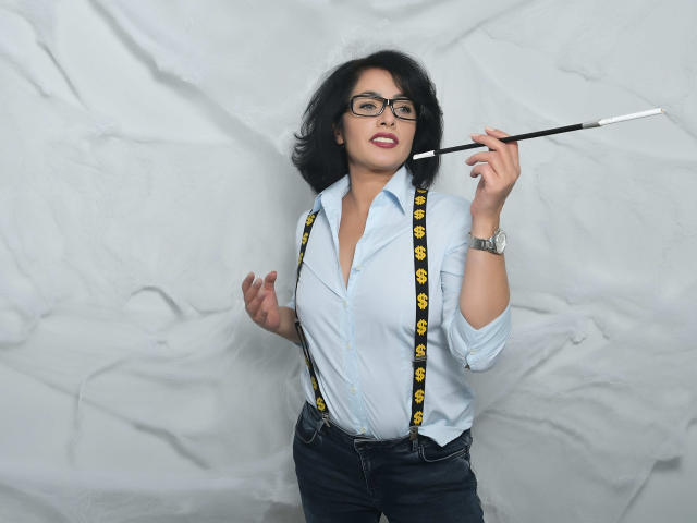 AnelisaReys mit schwarzen Brillen, welligem, dunklem Haar, einem blauen Hemd, gelben, mit einem Smiley-Gesicht versehenen Hosenträgern, dunklen Jeans, die einen Bleistift hält, lächelnd, mit einer silbernen Uhr am Handgelenk, vor einem weißen Hintergrund..