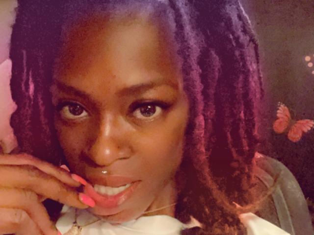 PrettyPrisca, con rastas moradas, un piercing en la nariz, uñas rosas y una ligera sonrisa, mirando directamente a la cámara con una expresión linda y sexy. Su piel marrón oscuro es suave..