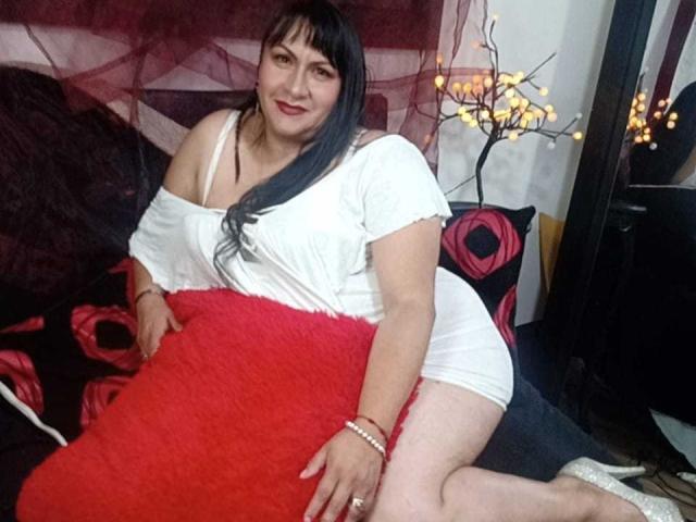 LauraMimiHot, formosa con capelli neri e rossetto rosso, indossa un abito bianco con una spalla scoperta, seduta su un letto nero con cuscini rossi, tenendo un cuscino rosso soffice a forma di seno..