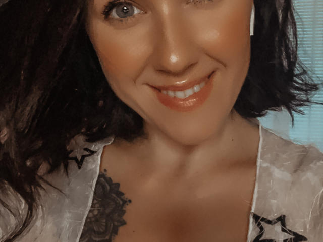 Misstyk with wavy dark hair, glossy orange lipstick, and a tattoo on her right breast, wearing a sheer white top, smiling at the camera. Her tits are slightly visible.