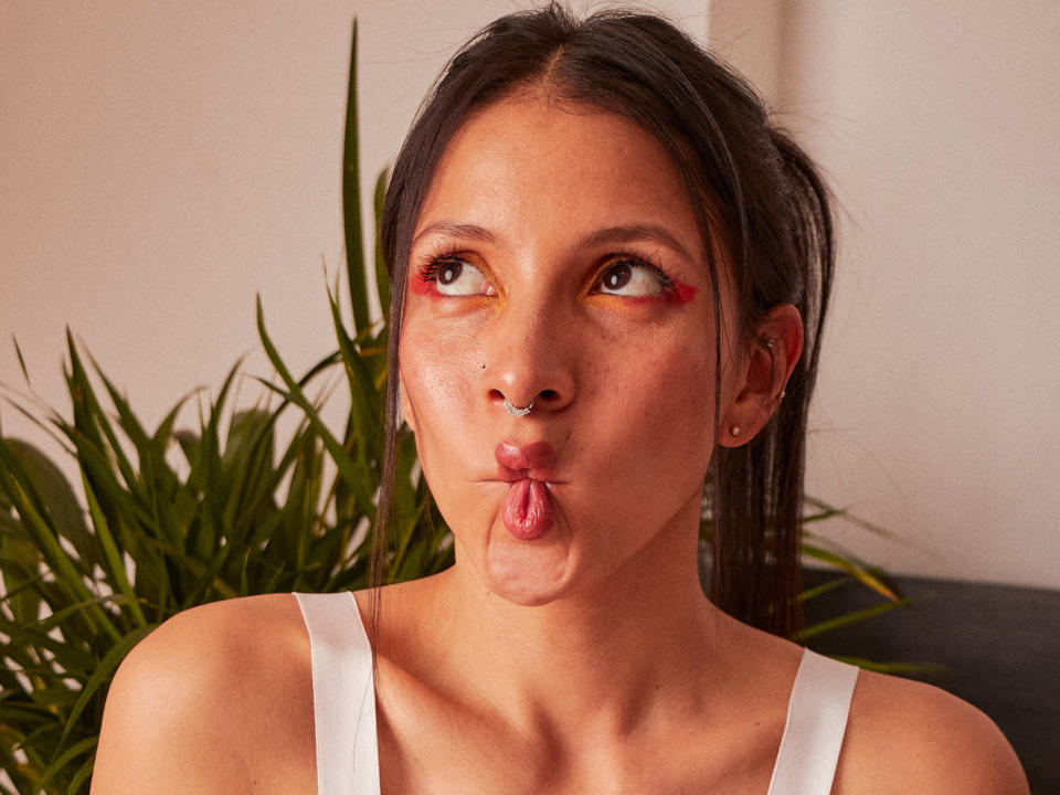 Julieth Vega, con labios fruncidos, aro en la nariz, cabello oscuro en una coleta, camiseta blanca, maquillaje de ojos rojo y amarillo, una planta verde en el fondo, pequeños aretes y una expresión facial linda..