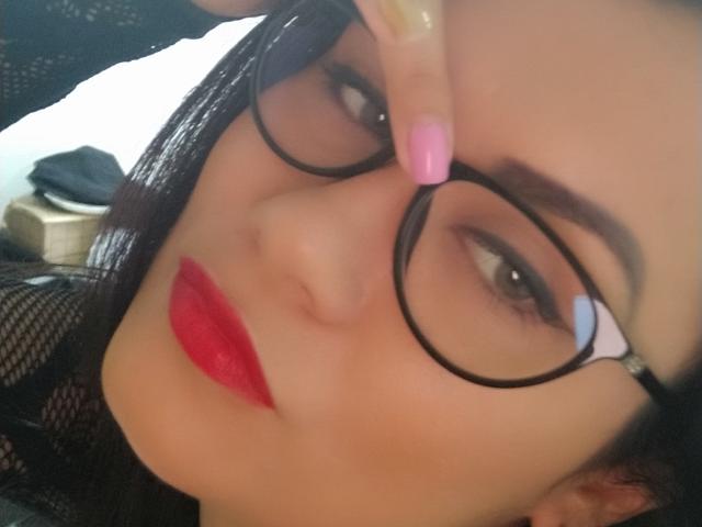 XBellaEvaX with red lipstick, black glasses, and pink nail polish, touching her glasses with her finger. She has dark hair and a slightly blurred background.