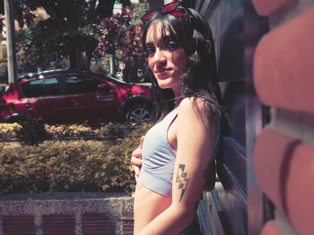 MeliMuller, con pequeños senos y un tatuaje en el brazo, se apoya contra una pared, vistiendo un top deportivo blanco y con gafas de sol en la cabeza, sonriendo a la luz del sol..