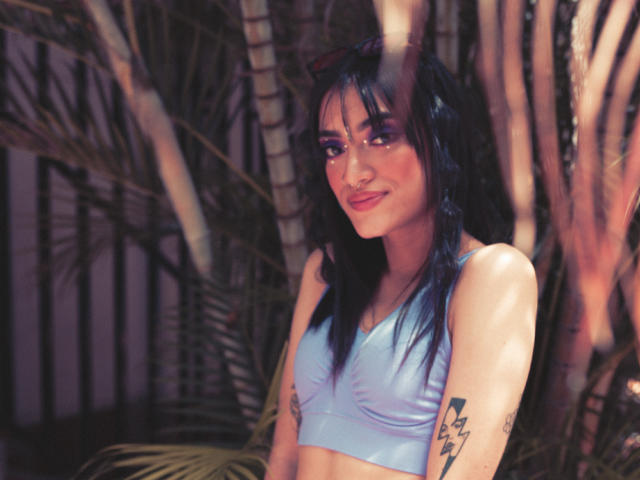 MeliMuller with blue hair, wearing a light blue sports bra, smiling, tattoos on arms, standing among palm trees, sunlight filtering through leaves.