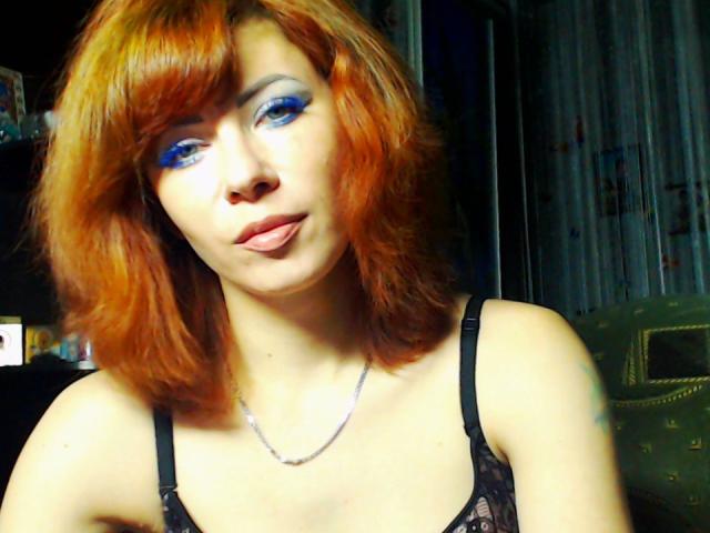 JuliaIce, con i capelli rossi, il trucco blu sugli occhi e le labbra carnose, indossa una camicetta di pizzo nero che mette in mostra il seno, e una collana d'argento, appare carina e sexy..
