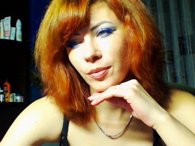 JuliaIce, avec des cheveux roux, du maquillage bleu pour les yeux, la langue tirée, une main sous le menton, portant un haut noir et un collier en chaîne. Seins visibles, peau pâle, léger sourire..