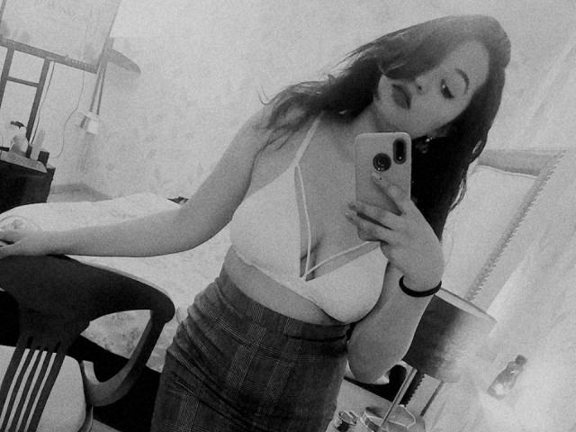 IsabellaWang met borsten, in een witte bh, een geruite rok en lang donker haar, die een selfie maakt in een spiegel met een telefoon, leunend op een stoel..