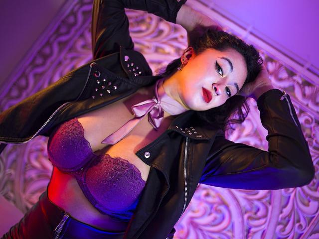 Isabella Wang, com cabelo escuro, vestindo uma jaqueta de couro preta, um sutiã de renda roxo e um colar de fita roxa, com os braços levantados, iluminação roxa e vermelha, mostrando seus seios grandes e seu rosto sensual..