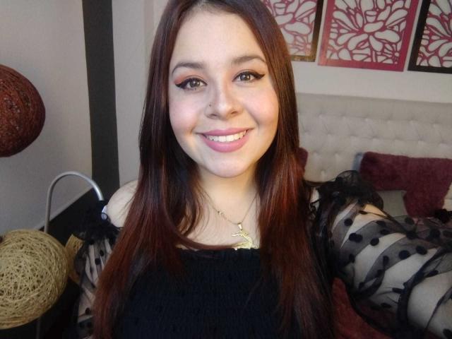 Katie Woolf, avec de longs cheveux bruns, souriante, portant un haut noir avec des manches transparentes à pois, un collier en or et du maquillage rose pour les yeux, montrant ses seins dans une chambre..