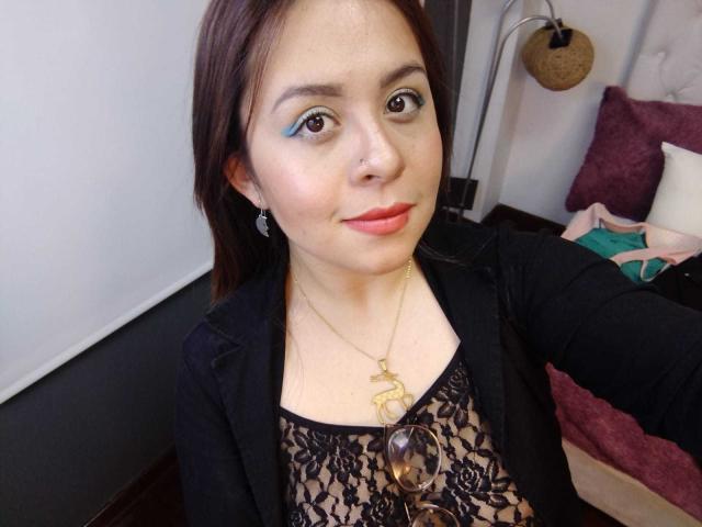 Katie Woolf, met bruine ogen, draagt een zwarte top met kant, een zwarte jas, een gouden ketting en rode lippenstift, ze glimlacht licht, heeft bruin haar en een kleine neusring, en houdt een zonnebril vast..