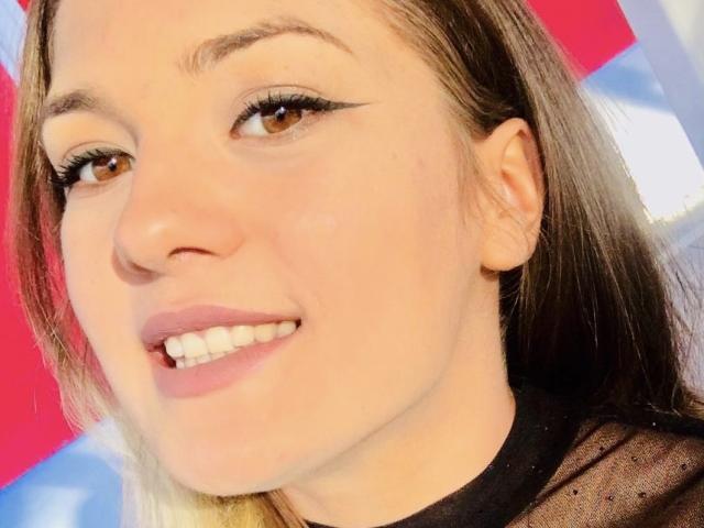 Le visage de SweetJanie, avec ses yeux marron, son eye-liner noir et son sourire où l'on voit ses dents. Elle porte un haut noir transparent, et ses cheveux bruns clairs sont coiffés avec une raie..