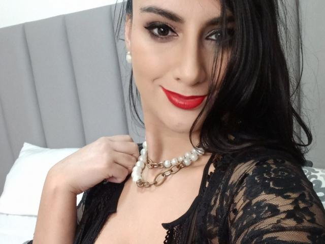 Julia Robinson usando uma blusa preta de renda, batom vermelho, colar de pérolas e uma corrente, segurando a blusa, sorrindo, mostrando os seios, cabelo preto e um travesseiro branco ao fundo..