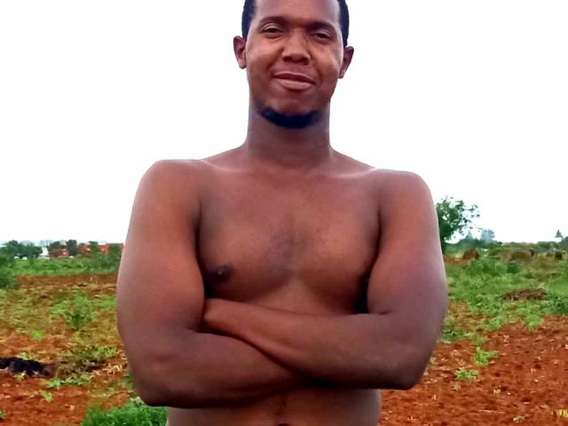 Milakay é um homem de pele escura, sem camisa, com uma pequena barba, braços cruzados e um leve sorriso, em pé em uma área rural suja e verde..