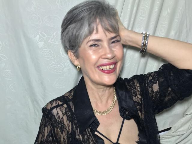 BelleBlondx with short gray hair, black lace top, gold necklace, and pink lipstick, smiling, hand in hair, wearing bracelets, looking cute and confident.