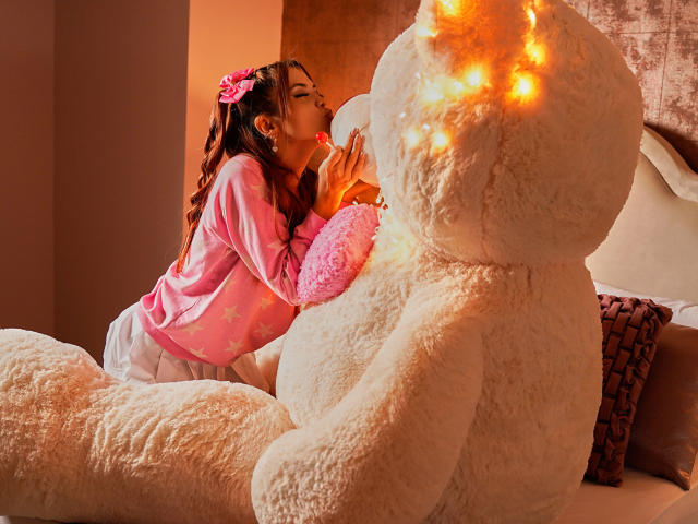 Maisy Warner, avec ses cheveux roses, embrasse un ours en peluche géant et moelleux de couleur blanche, elle porte un haut rose à manches longues et un short blanc, avec du rouge à lèvres rose sur ses lèvres. Une lumière chaude vient d'en haut..
