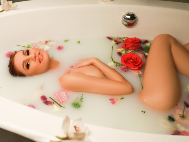 Julia Richard en una bañera blanca llena de agua y pétalos de flores rosas, blancos y rojos, acostada desnuda con los senos cubiertos, sonriendo a la cámara, con las piernas cruzadas..
