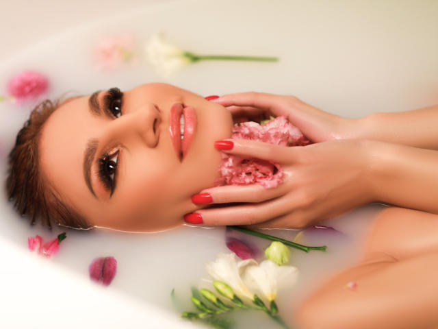 Le visage de Julia Richard dans une baignoire blanche, tenant des pétales de rose roses près de sa bouche avec ses ongles rouges. Ses yeux marrons sont à moitié ouverts, et elle a des lèvres roses et brillantes..