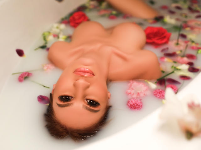 JuliaRichard, nua, com os seios acima da água, deitada em uma banheira branca cheia de pétalas de rosa e água, olhando para cima com um sorriso suave, olhos castanhos e lábios rosados..