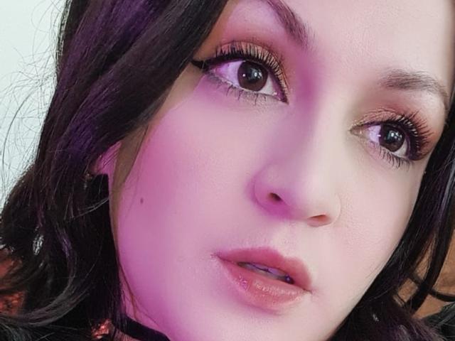 ElinaJhonson's face with purple lighting, showing her brown eyes, long eyelashes, pinkish lips, and dark wavy hair, looking slightly to the side with a neutral expression.
