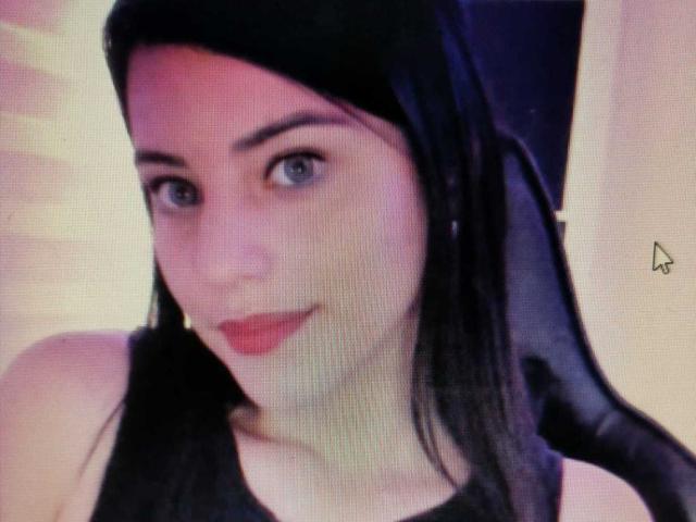 Arenne with pale skin, black hair, and blue eyes, wearing a black headscarf, smiling with red lipstick, showing her tits slightly in a black tank top.