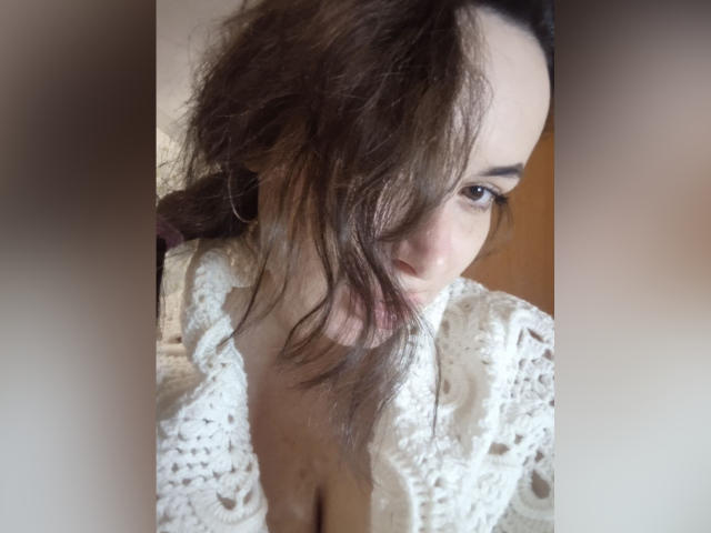 LarisaMons met rommelig bruin haar, die zijwaarts kijkt, gekleed in een witte gebreide top die haar decolleté laat zien, met bruine ogen en kleine oorringen, op een zacht verlichte, licht onscherpe foto..