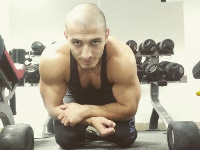 AdamTasty, kaal, gespierd, zwart tanktop, knielend, armen gekruist, naar de camera kijkend, sportschool, zwarte haltergewichten, serieuze uitdrukking, bezweet, getraind, geconcentreerd, donkere broek, vloerbedekt met tapijt..