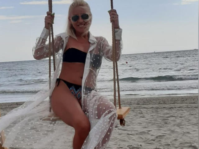 Mary, con i capelli biondi, che indossa occhiali da sole, un abito trasparente a pois, un bikini nero, seduta su un'altalena di legno, che tiene le corde, sorridente, spiaggia, oceano, piccole onde, cielo nuvoloso..