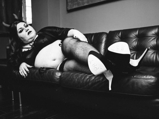 KaryQueen lounges on a leather couch, legs up, showing her bare ass and hairy legs. She wears black heels, a choker, and a sheer top. Black-and-white photo.