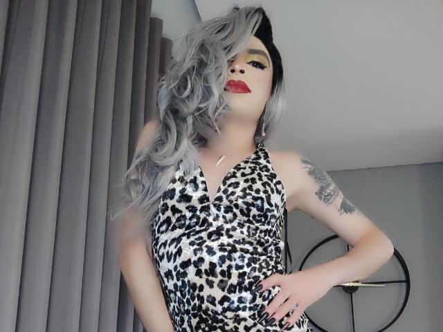 Rosariio met lang grijs haar, een jurk met luipaardprint, rode lippenstift, gele oogschaduw, zwarte kattenoren, een tatoeage op de rechterarm, staand met een hand op de heup, grijze gordijnen op de achtergrond..