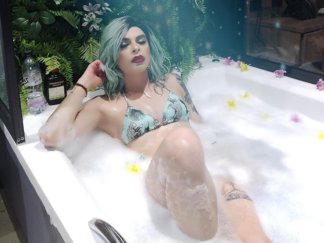 Rosariio met turquoise haar, die een groene, gestreepte bikini draagt, ontspant in een bubbelbad met wit water, met gekruiste benen, waarbij tatoeages zichtbaar zijn, met donkere lipstick, omringd door groen en gele bloemblaadjes..
