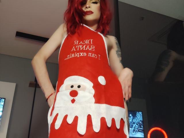 Rosariio mit roten Haaren, trägt ein rotes Weihnachtsmannkleid mit weißen Akzenten und einem Weihnachtsmanngesicht, steht mit den Händen in den Hüften und zeigt ein Tattoo am rechten Arm..