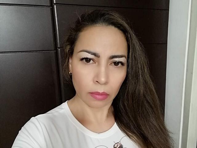 A SofyGray tem cabelos castanhos longos, olhos castanhos, batom rosa e está vestindo uma camisa branca com um pequeno desenho vermelho. Suas sobrancelhas estão bem definidas..