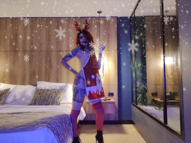 Rosariio stands in a bedroom wearing a Santa dress with red and white fur trim, red thigh-high socks, and reindeer antlers. She's holding a snowflake, showing tattoos.