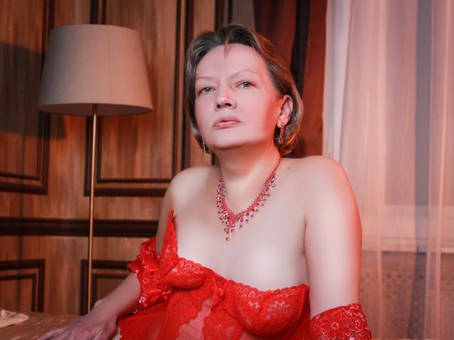 GranyLoveSex with short brown hair, red lace top, exposed boobs, red necklace, earrings, and a confident expression in a warmly lit room. Lamp and curtain in background.