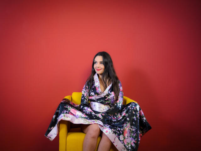 AshlyChloe, con cabello largo y oscuro, sentada en una silla amarilla, vestida con un kimono floral, mostrando sus muslos, y sonriendo con lápiz labial rojo, frente a una pared roja..