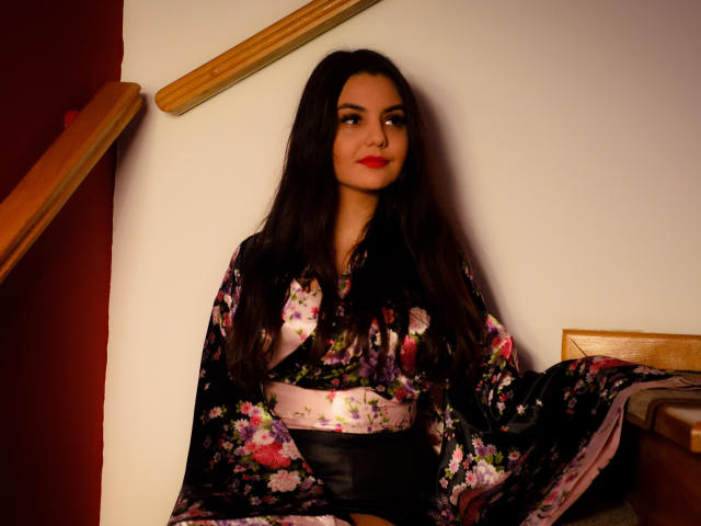 AshlyChloe con lunghi capelli scuri, rossetto rosso, un kimono nero con fiori, una cintura bianca, una cintura nera, seduta su gradini di legno, che guarda leggermente di lato con un sorriso appena accennato..
