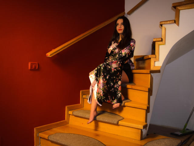 AshlyChloe sits on wooden stairs with a floral dress, bare feet, and a wooden handrail. She has dark hair, a red wall, and a white wall behind her.