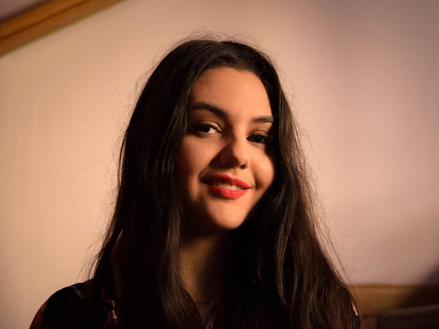 AshlyChloe has long, dark, slightly messy hair and a soft smile with red lips. She's looking to the side with warm, subtle lighting on her face.