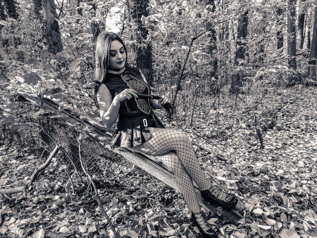 AshlyChloe sitting on broken wooden chair in forest, wearing fishnet stockings, black top, and skirt, holding shotgun, with pale skin, blonde hair, and black lipstick.
