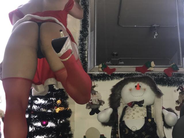 BellaLolita's ass in a red dress with black thong, red stockings, and high heels, next to a Christmas tree and a stuffed snowman in a decorated room.